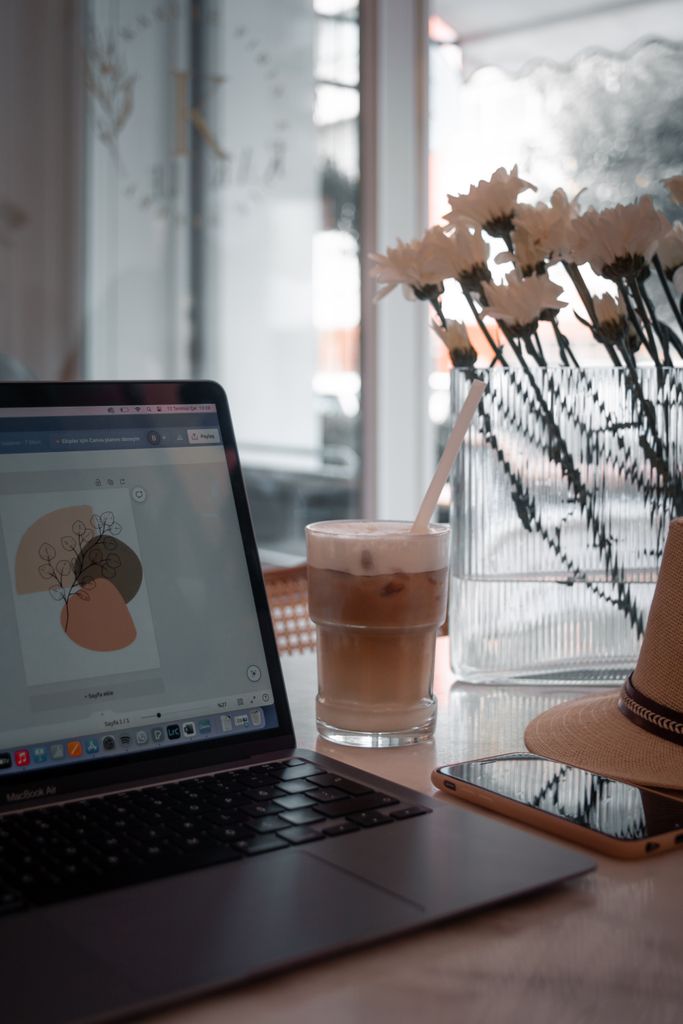 A relaxing café scene featuring a laptop, iced coffee, flowers, and a hat by the window.